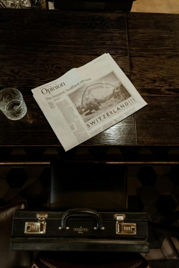 Top view of a newspaper and glass beside a briefcase on a vintage wooden table.